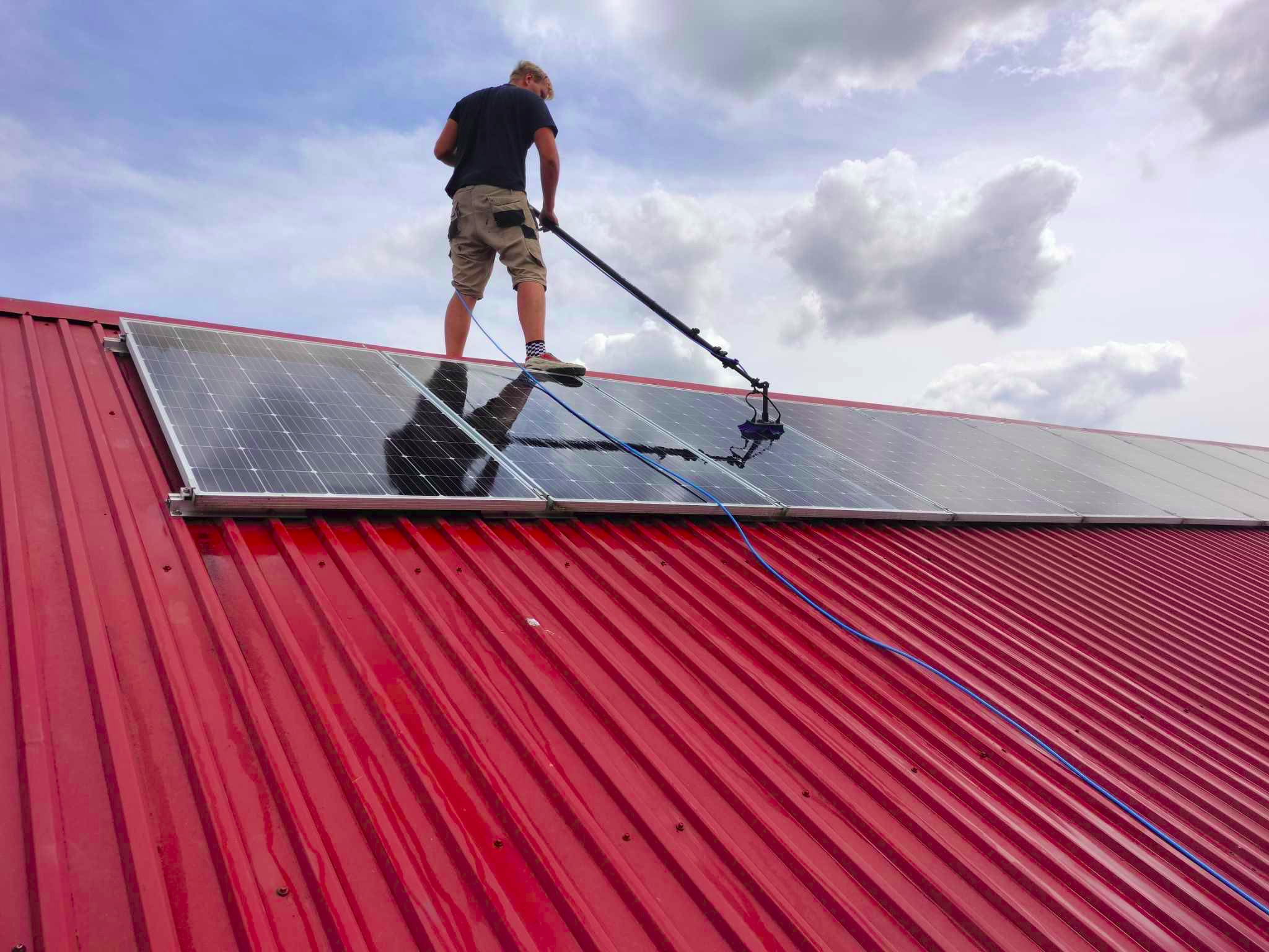 Mężczyzna w krótkich spodenkach i skarpetkach w paski czyści panele słoneczne na czerwonym blaszanym dachu za pomocą teleskopowej lancy z szczotką, widoczne niebo z chmurami.
