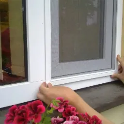 Montaż ramkowej siatki okiennej na białym oknie PCV, widoczne dłonie montera i doniczka z czerwonymi kwiatami na parapecie.