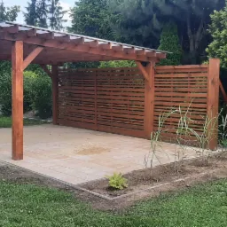 Drewniana pergola z zadaszeniem i ażurową ścianą, zbudowana na świeżo ułożonym placu z jasnych płyt brukowych, otoczona zielenią ogrodową i młodymi roślinami wzdłuż krawędzi.