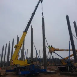 Żółty dźwig z napisem 'KRANTRANS' podnosi betonowy słup na placu budowy; w tle widoczne liczne słupy i podnośnik koszowy z pracownikami w kamizelkach odblaskowych.