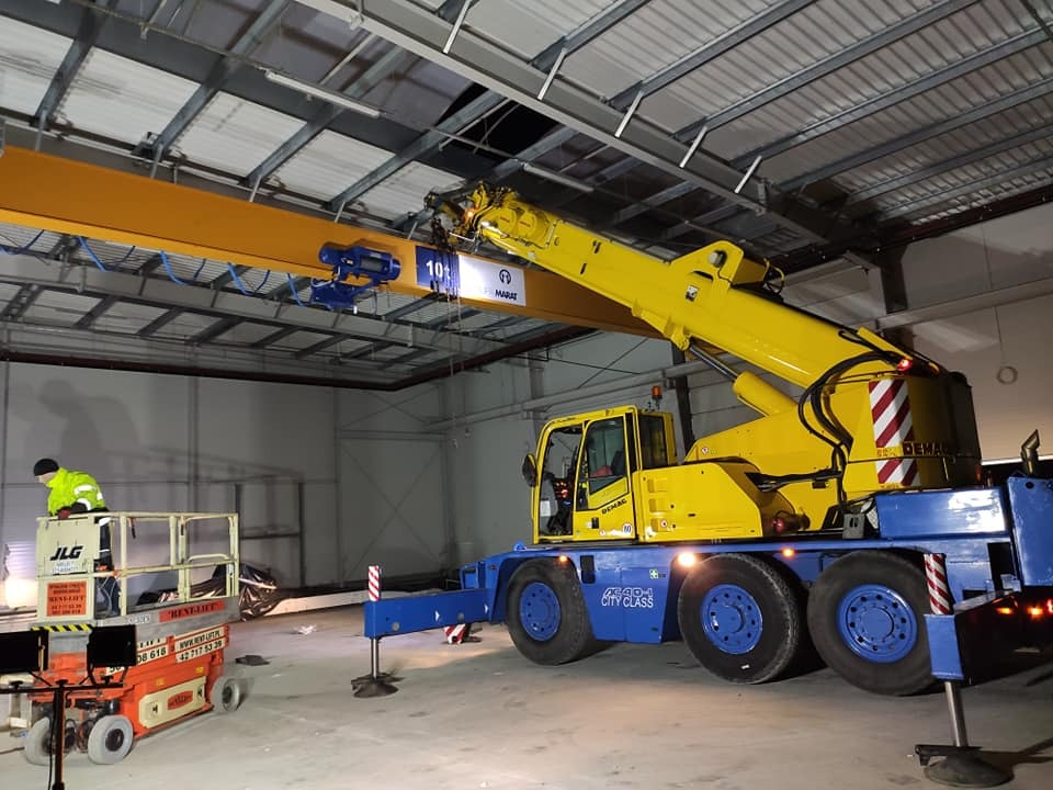 Żółto-niebieski dźwig samochodowy Demag City Class w hali, z wysięgnikiem pod stropem, obok podnośnik nożycowy JLG z pracownikiem w odblaskowej kurtce.