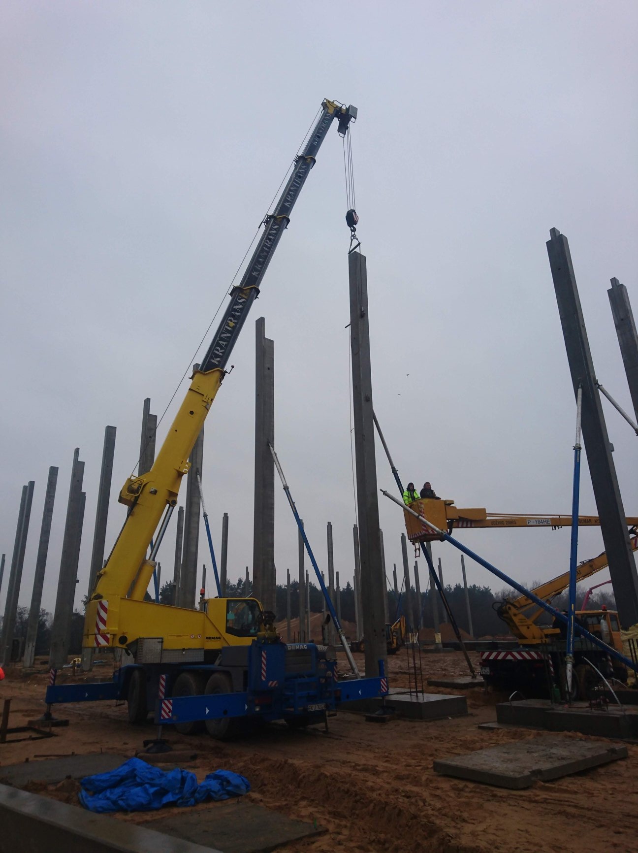 Żółty dźwig z napisem 'KRANTRANS' podnosi betonowy słup na placu budowy; w tle widoczne liczne słupy i podnośnik koszowy z pracownikami w kamizelkach odblaskowych.