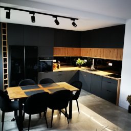 LukArt Łukasz Linczowski - Nowoczesna kuchnia w Rzeszowie z czarnymi matowymi szafkami, drewnianym blatem i stołem z czarnymi krzesłami. Oświetlenie podszafkowe podkreśla naturalne drewno.