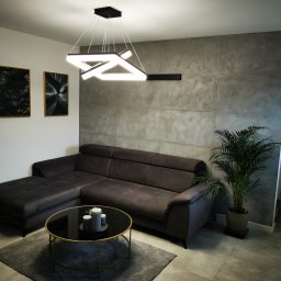 LukArt Łukasz Linczowski - Szary narożnik z poduszkami na tle ściany wykończonej płytami imitującymi beton, okrągły stolik kawowy z czarnym blatem i złotą ramą, nowoczesna lampa wisząca LED o geometrycznym kształcie, dywan...