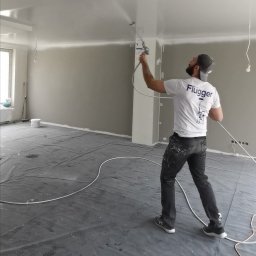 LukArt Łukasz Linczowski - Mężczyzna w czapce i koszulce Flűgger maluje sufit natryskowo w pomieszczeniu zabezpieczonym folią, widoczne ślady farby na spodniach i podłodze.