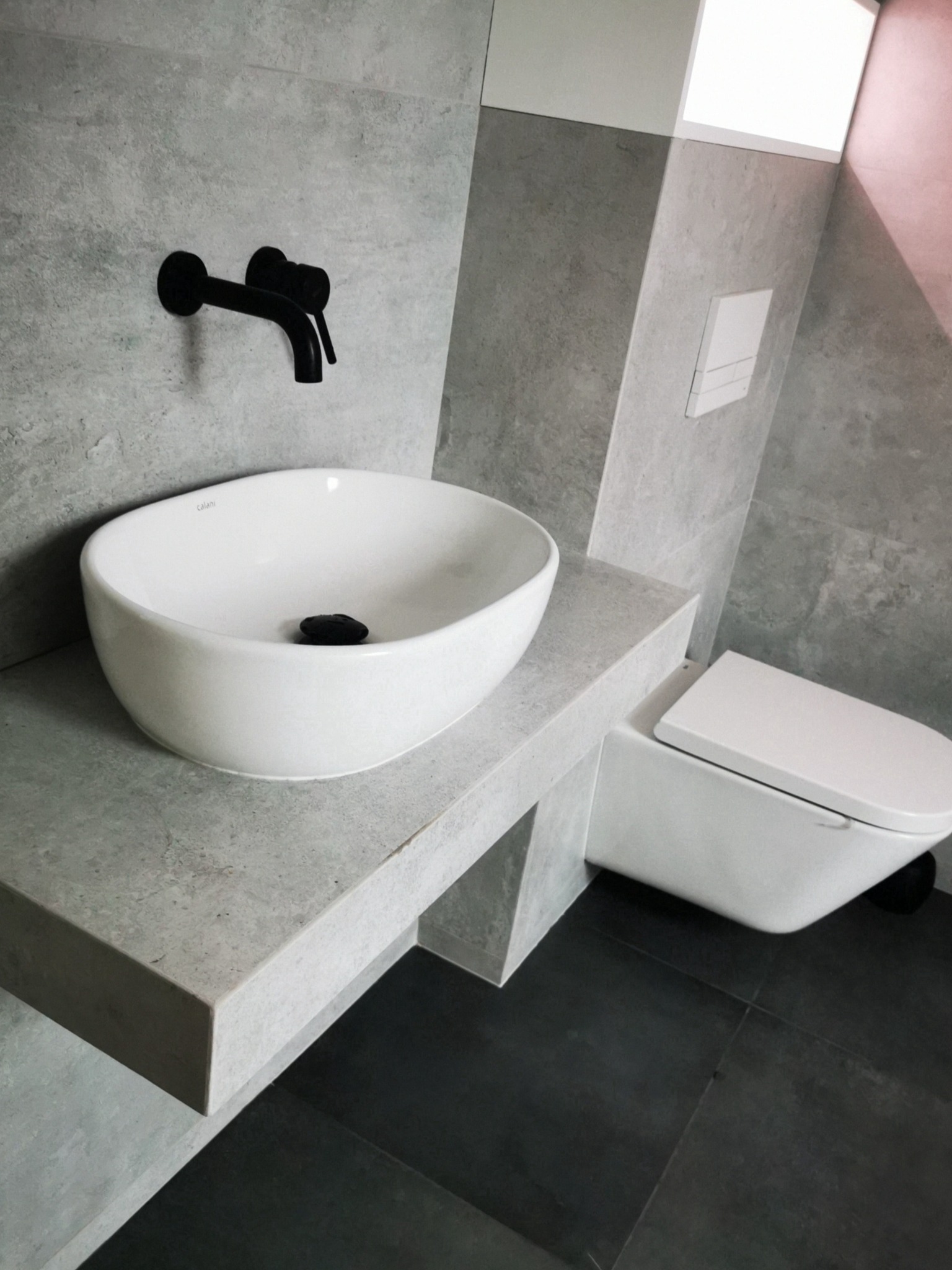 Nowoczesna łazienka z owalną białą umywalką nablatową, czarną armaturą i podwieszaną toaletą w szaro-białej kolorystyce.