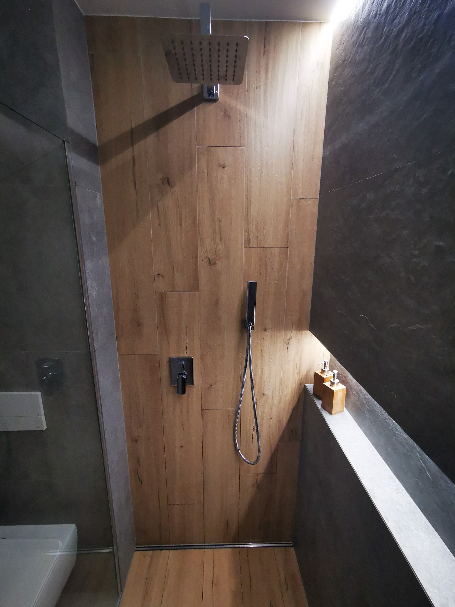 Nowoczesna kabina prysznicowa z drewnopodobnymi płytkami na ścianie, deszczownicą i czarną armaturą, uzupełniona o dozowniki mydła na półce.