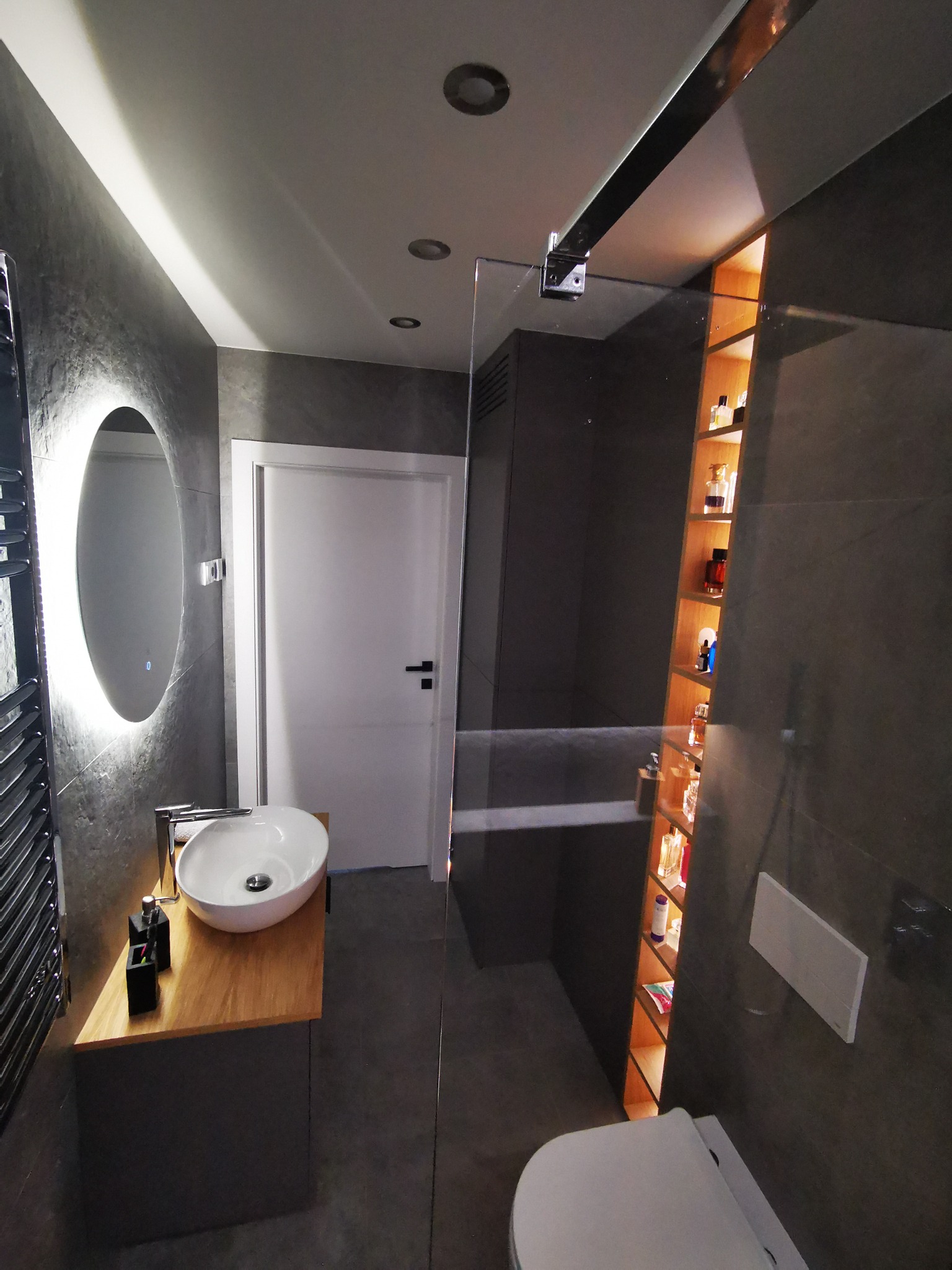 Nowoczesna łazienka z szarymi płytkami, owalnym lustrem z podświetleniem LED, drewnianym blatem pod umywalką nablatową i przeszklonym prysznicem z półkami na kosmetyki.