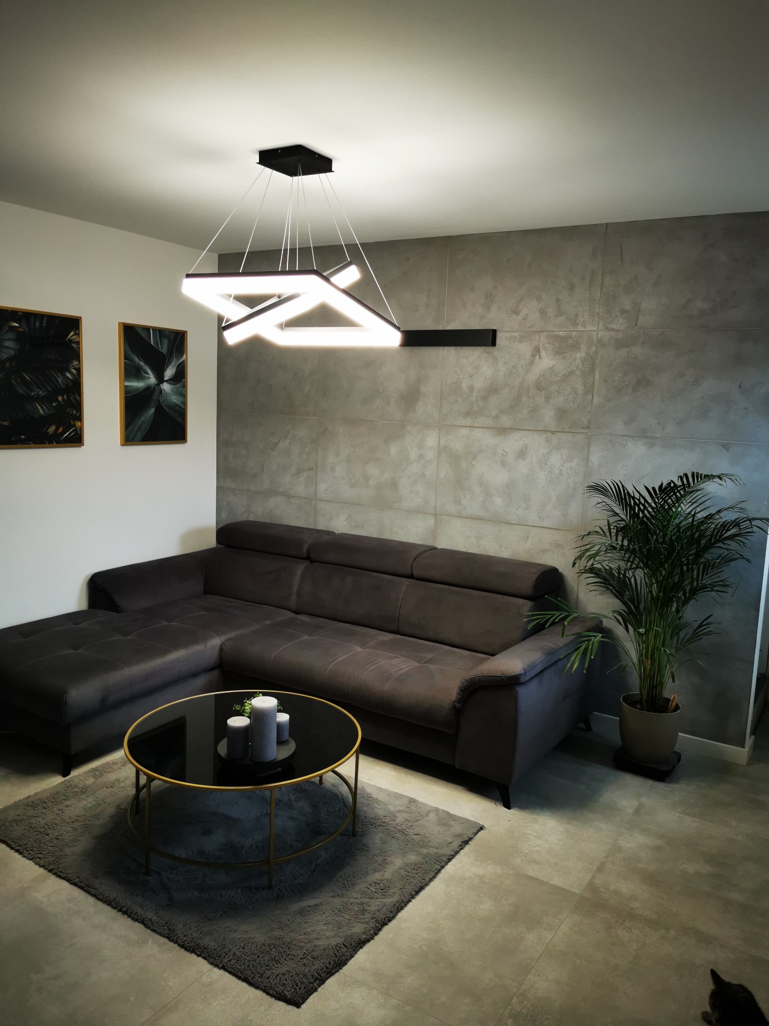 Szary narożnik z poduszkami na tle ściany wykończonej płytami imitującymi beton, okrągły stolik kawowy z czarnym blatem i złotą ramą, nowoczesna lampa wisząca LED o geometrycznym kształcie, dywan...