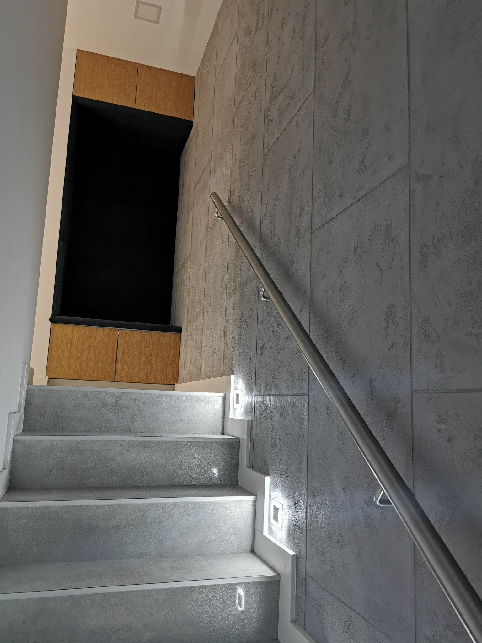 Schody z szarymi stopniami, podświetleniem LED w ścianie i metalową poręczą, obok wnęka z drewnianymi szafkami.