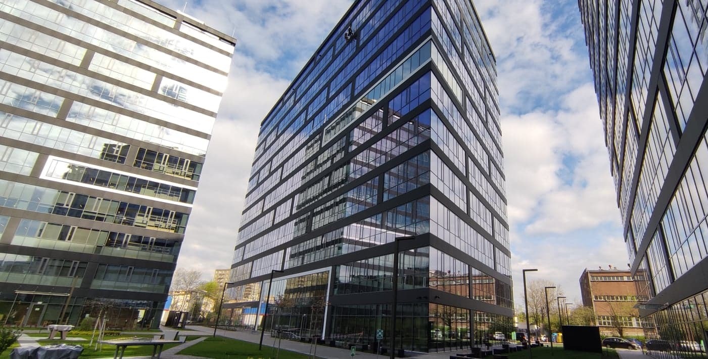 Nowoczesny biurowiec z lustrzanymi fasadami odbijającymi błękitne niebo z chmurami, widok z poziomu trawnika z ławkami.