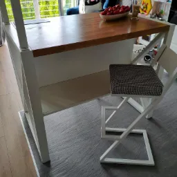 Wysoki stół barowy z drewnianym blatem i białymi nogami, stojący na szarej podłodze, z białym krzesłem o geometrycznym wzorze na siedzisku, w tle okno i kolorowa ściana z panelami.