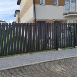 Nowoczesna, grafitowa brama wjazdowa z pionowych sztachet, z czarną skrzynką na listy, na tle dwukondygnacyjnego domu z balkonem.