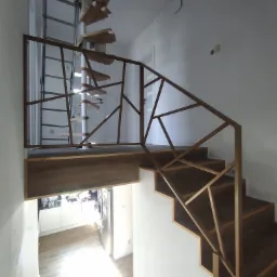 Nowoczesna balustrada o geometrycznym wzorze, wykonana z metalu w kolorze starego złota, zamontowana przy drewnianych schodach w jasnym wnętrzu, z widoczną spiralną klatką schodową w tle.