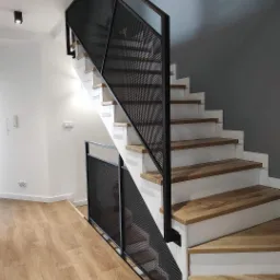 Nowoczesne schody drewniane z białymi podstopnicami i czarną, metalową balustradą z siatki, widok z dołu na jasne wnętrze z parkietem i szarą ścianą w tle.