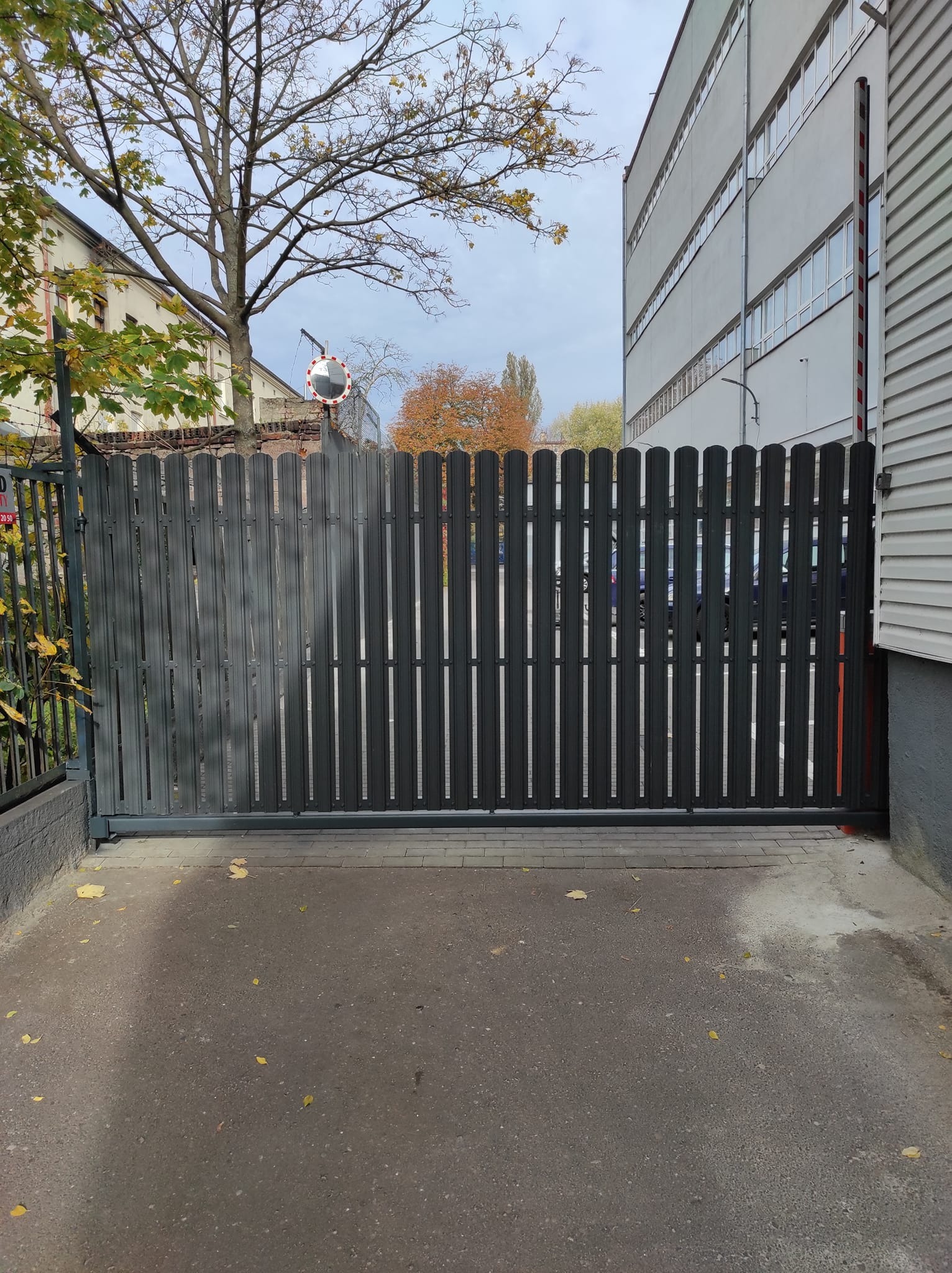 Zasuwana, metalowa brama wjazdowa z pionowych sztachet w kolorze grafitowym, zamontowana przed budynkiem, z widocznym fragmentem chodnika i opadłymi liśćmi.