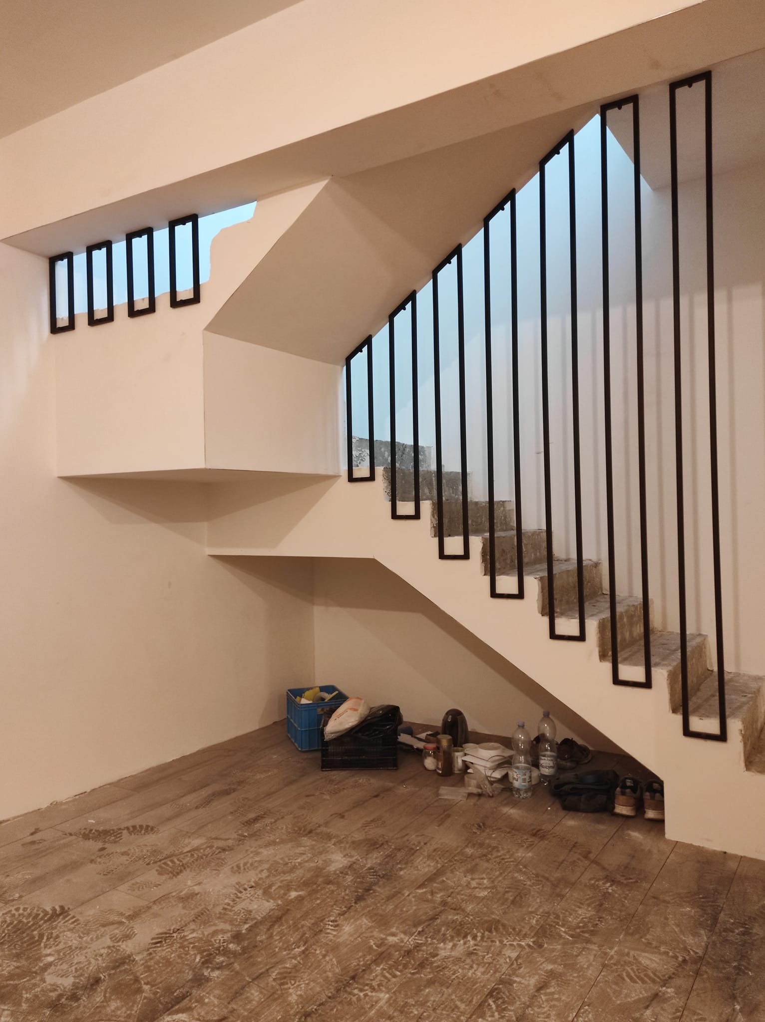 Wnętrze w trakcie remontu: betonowe schody z czarnymi, prostymi balustradami, podłoga pokryta śladami butów, widoczne narzędzia i materiały budowlane.