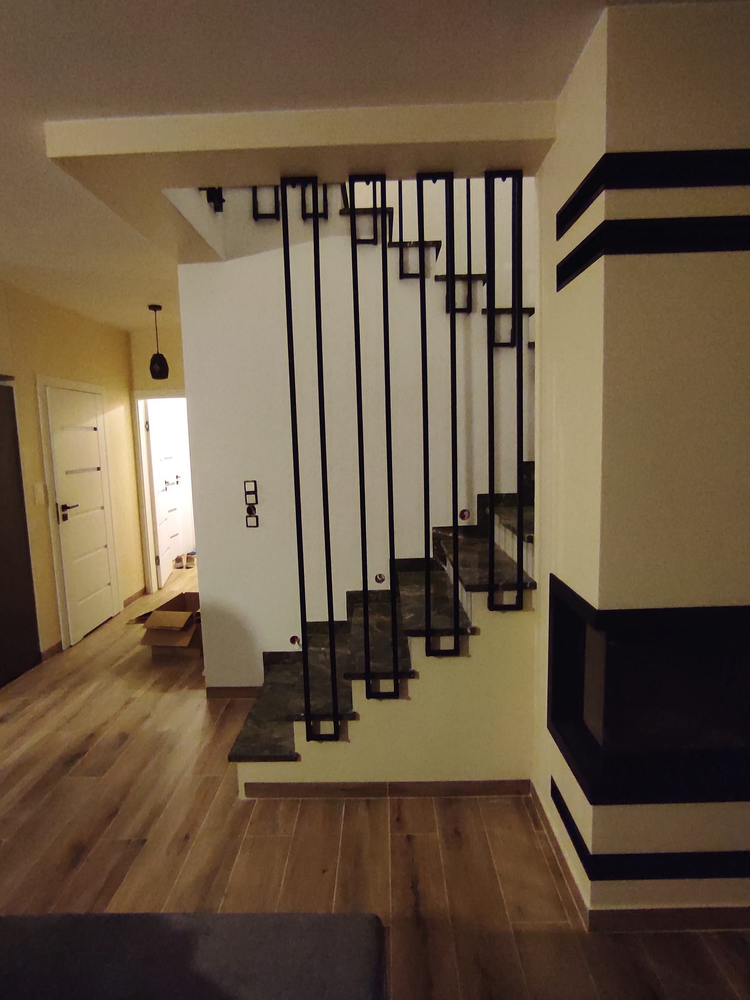 Nowoczesne schody z czarnymi metalowymi balustradami o geometrycznym wzorze, widoczne z dołu, z fragmentem kominka po prawej stronie i otwartym przejściem do innego pomieszczenia w tle.