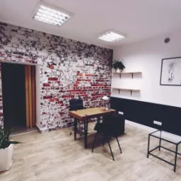 Nowoczesne biuro z drewnianym biurkiem, czarnymi krzesłami i ścianą z cegły pomalowanej na biało, oświetlone lampami sufitowymi. Na ścianach wiszą grafiki w czarnych ramkach.