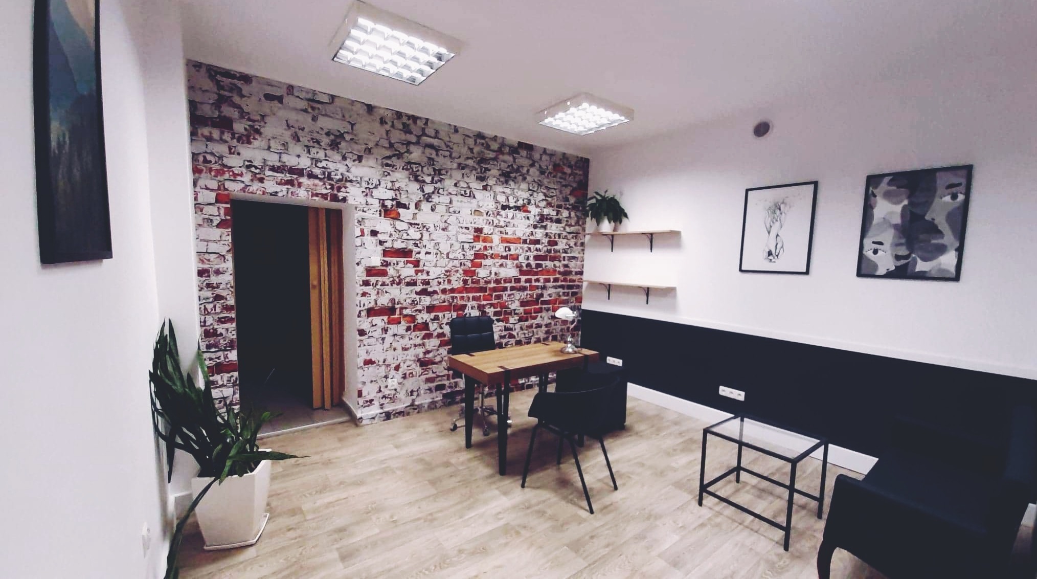 Nowoczesne biuro z drewnianym biurkiem, czarnymi krzesłami i ścianą z cegły pomalowanej na biało, oświetlone lampami sufitowymi. Na ścianach wiszą grafiki w czarnych ramkach.