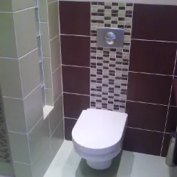 Wykończona toaleta z podwieszaną muszlą, brązowymi i mozaikowymi płytkami oraz chromowanym przyciskiem spłukiwania, widoczna z poziomu podłogi.