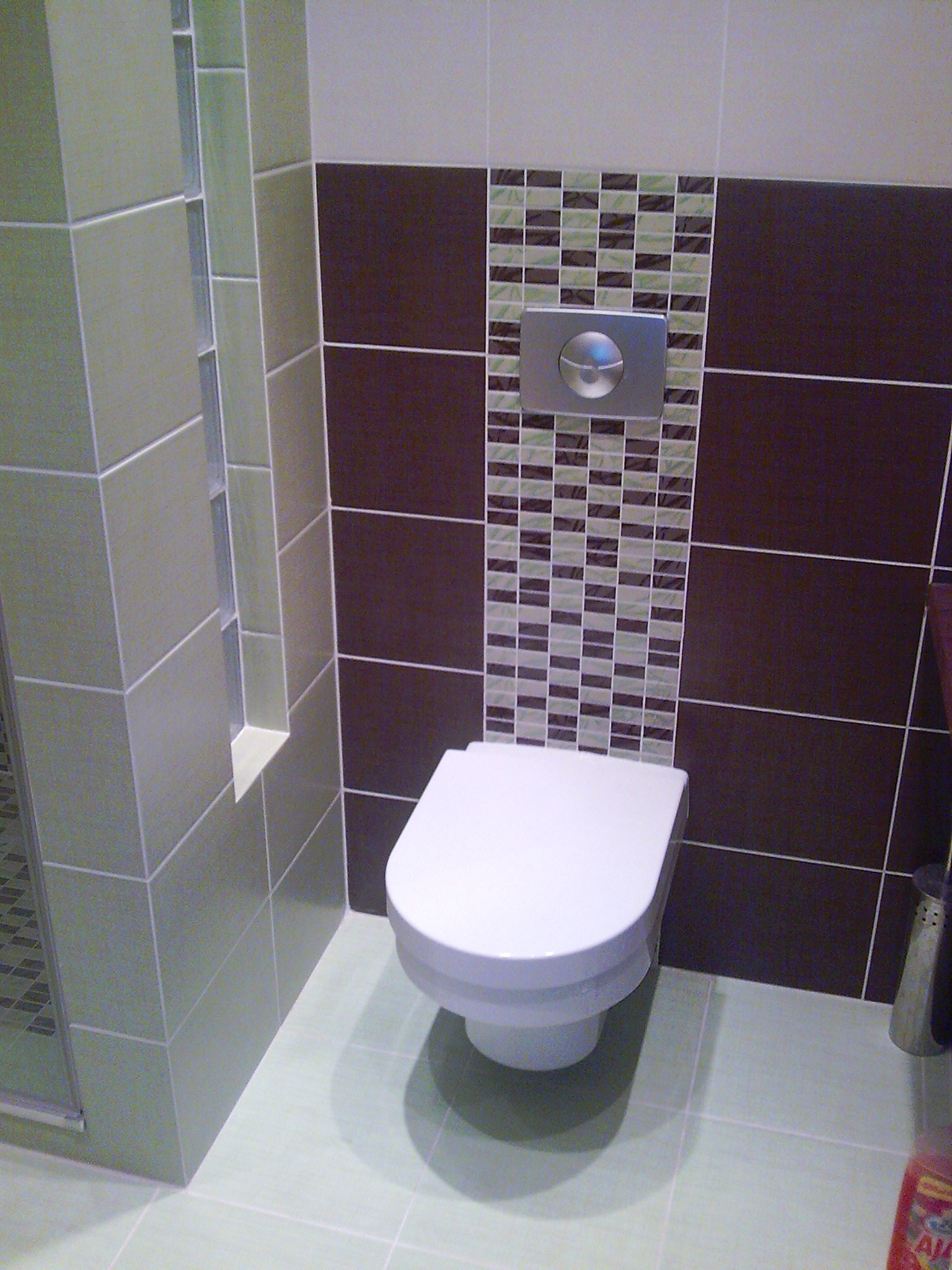 Wykończona toaleta z podwieszaną muszlą, brązowymi i mozaikowymi płytkami oraz chromowanym przyciskiem spłukiwania, widoczna z poziomu podłogi.