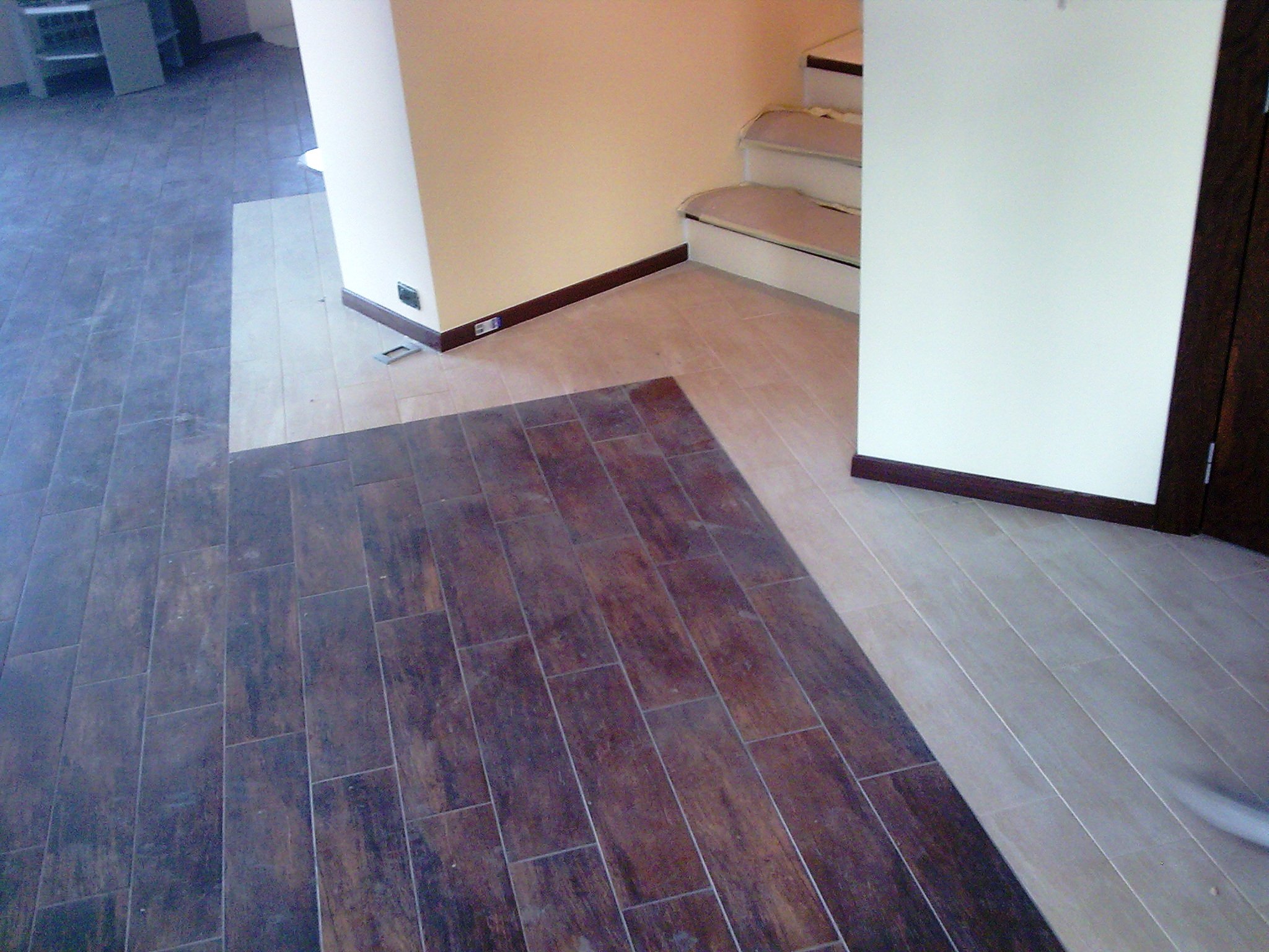 Układanie podłogi z płytek w dwóch odcieniach: ciemnobrązowym i beżowym, widoczne fragmenty schodów i ściany w jasnych kolorach, listwy przypodłogowe w kolorze ciemnego brązu.