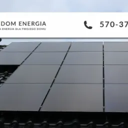 Panele fotowoltaiczne na czarnym dachu z dachówki ceramicznej, widok z dołu, logo Freedom Energia w górnej części zdjęcia.