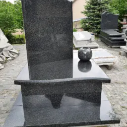 Czarny, granitowy nagrobek urnowy z zaokrągloną płytą główną i wazonem na górze, ustawiony na placu z kostki brukowej, z innymi nagrobkami w tle.