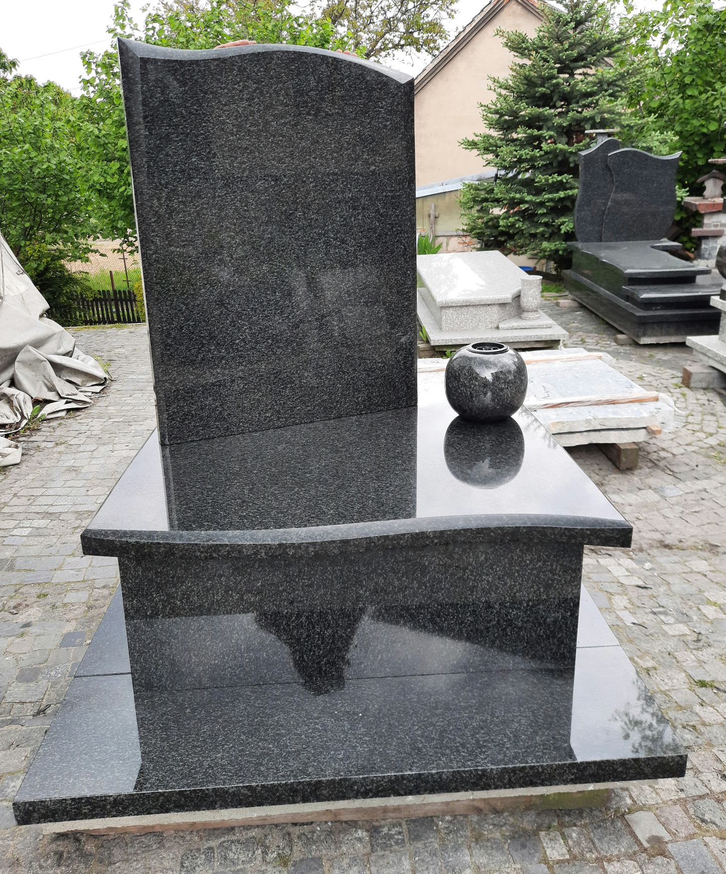 Czarny, granitowy nagrobek urnowy z zaokrągloną płytą główną i wazonem na górze, ustawiony na placu z kostki brukowej, z innymi nagrobkami w tle.