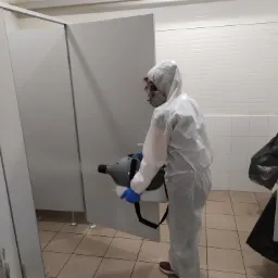 Pracownik w kombinezonie ochronnym i masce, przeprowadza odkażanie kabin toaletowych przy użyciu zamgławiacza.