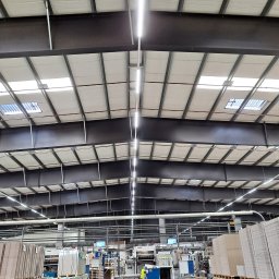 AR-TEX - Wnętrze hali produkcyjnej z widoczną konstrukcją dachu, oświetleniem LED i stosami gotowych produktów na paletach, pracownik w odblaskowej kamizelce obsługujący maszynę w tle.