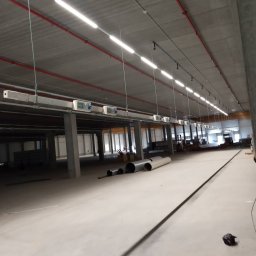 AR-TEX - Wnętrze hali przemysłowej w trakcie prac instalacyjnych: widoczne rury, kolumny wspierające, oświetlenie liniowe pod sufitem i instalacje elektryczne. Perspektywa z niskiego kąta ukazuje rozległość...