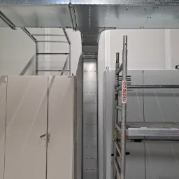 Metalowe koryto kablowe i drabinka zamontowane pod sufitem w pomieszczeniu technicznym, obok szafy elektrycznej i rusztowania z naklejką 'mateco'.