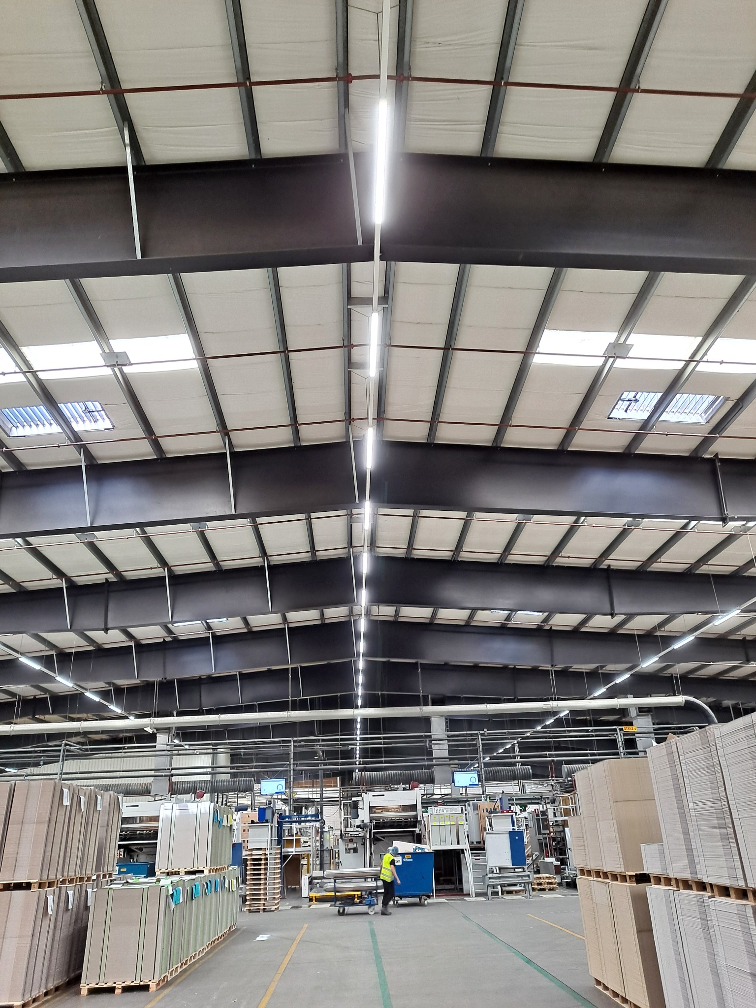 Wnętrze hali produkcyjnej z widoczną konstrukcją dachu, oświetleniem LED i stosami gotowych produktów na paletach, pracownik w odblaskowej kamizelce obsługujący maszynę w tle.
