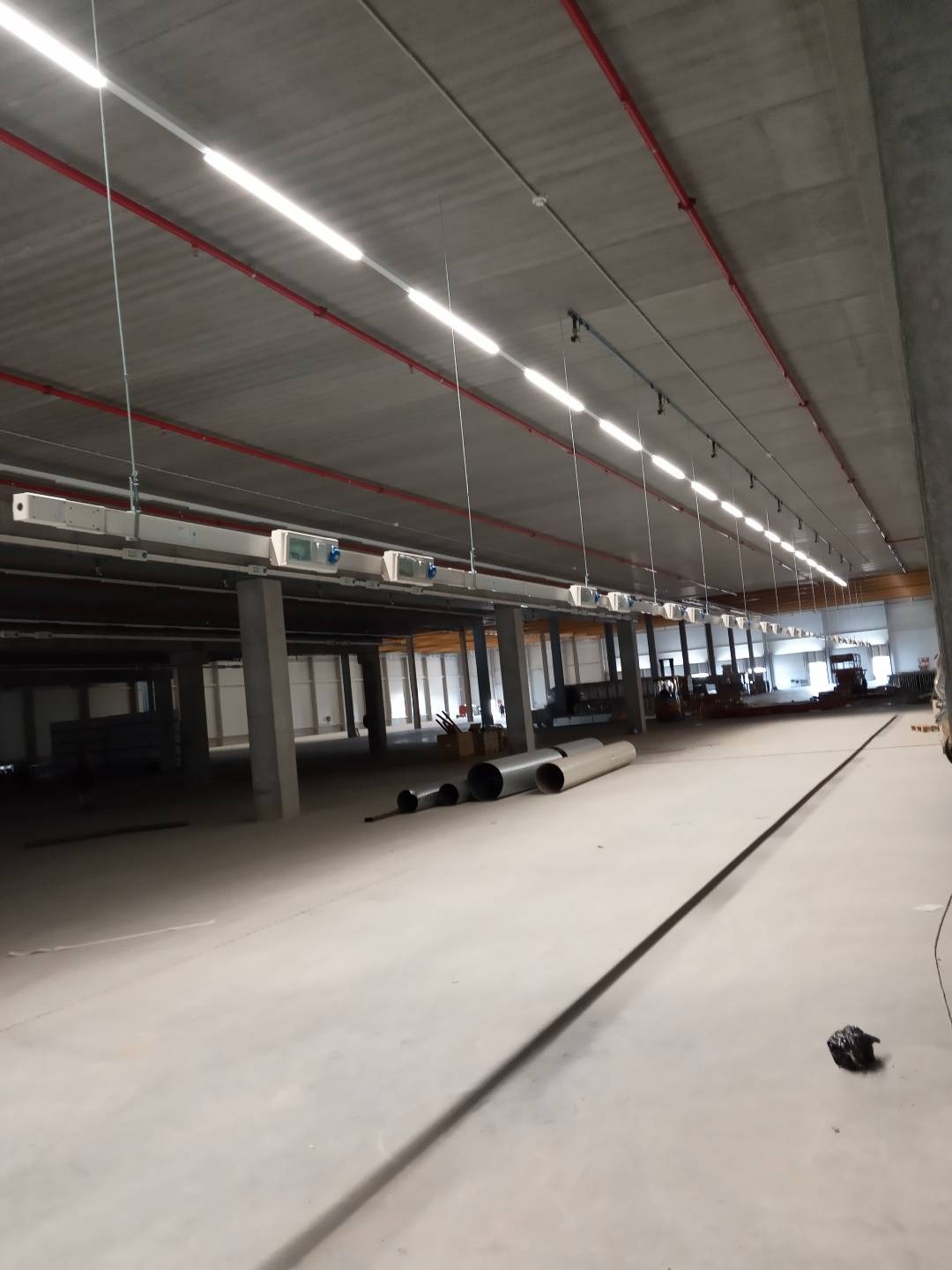 Wnętrze hali przemysłowej w trakcie prac instalacyjnych: widoczne rury, kolumny wspierające, oświetlenie liniowe pod sufitem i instalacje elektryczne. Perspektywa z niskiego kąta ukazuje rozległość...