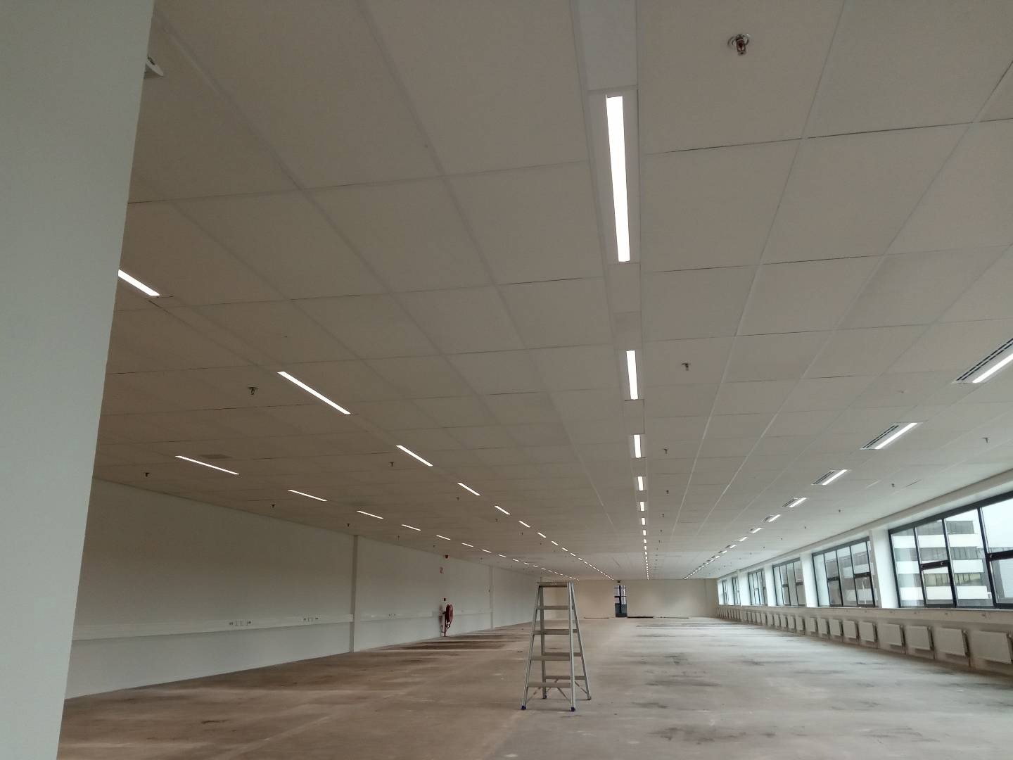 Rozległe wnętrze biurowe z rzędem długich, liniowych lamp LED w suficie podwieszanym, drabina aluminiowa po lewej stronie, duże okna wzdłuż ściany po prawej stronie.