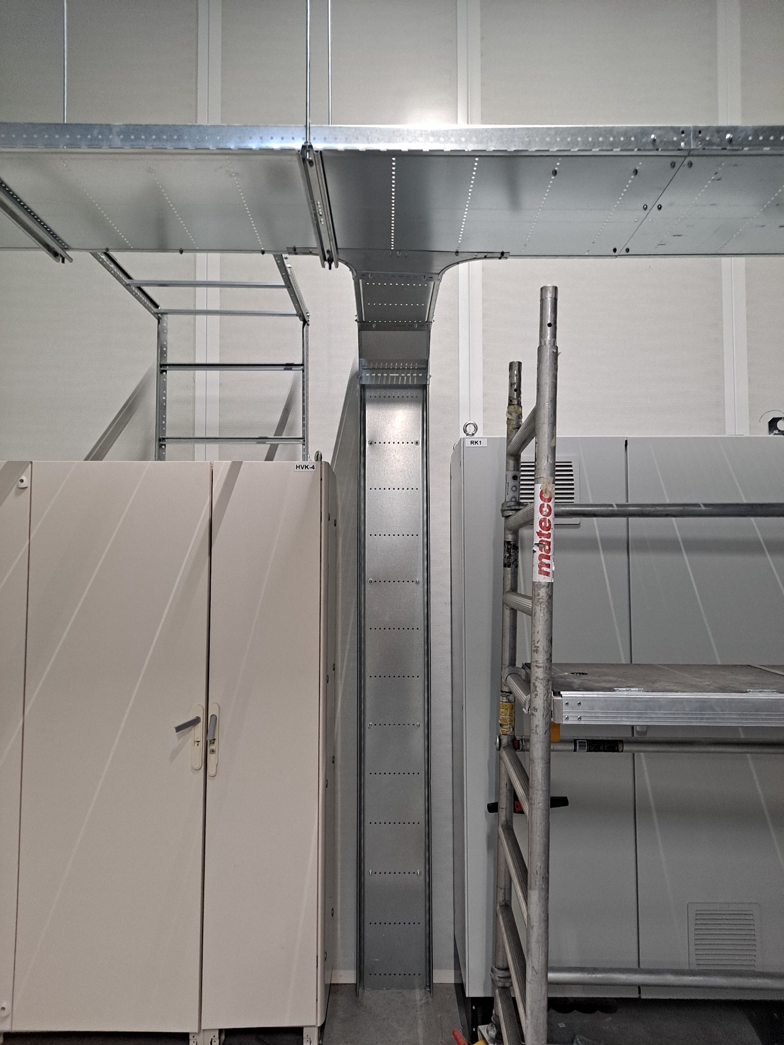 Metalowe koryto kablowe i drabinka zamontowane pod sufitem w pomieszczeniu technicznym, obok szafy elektrycznej i rusztowania z naklejką 'mateco'.