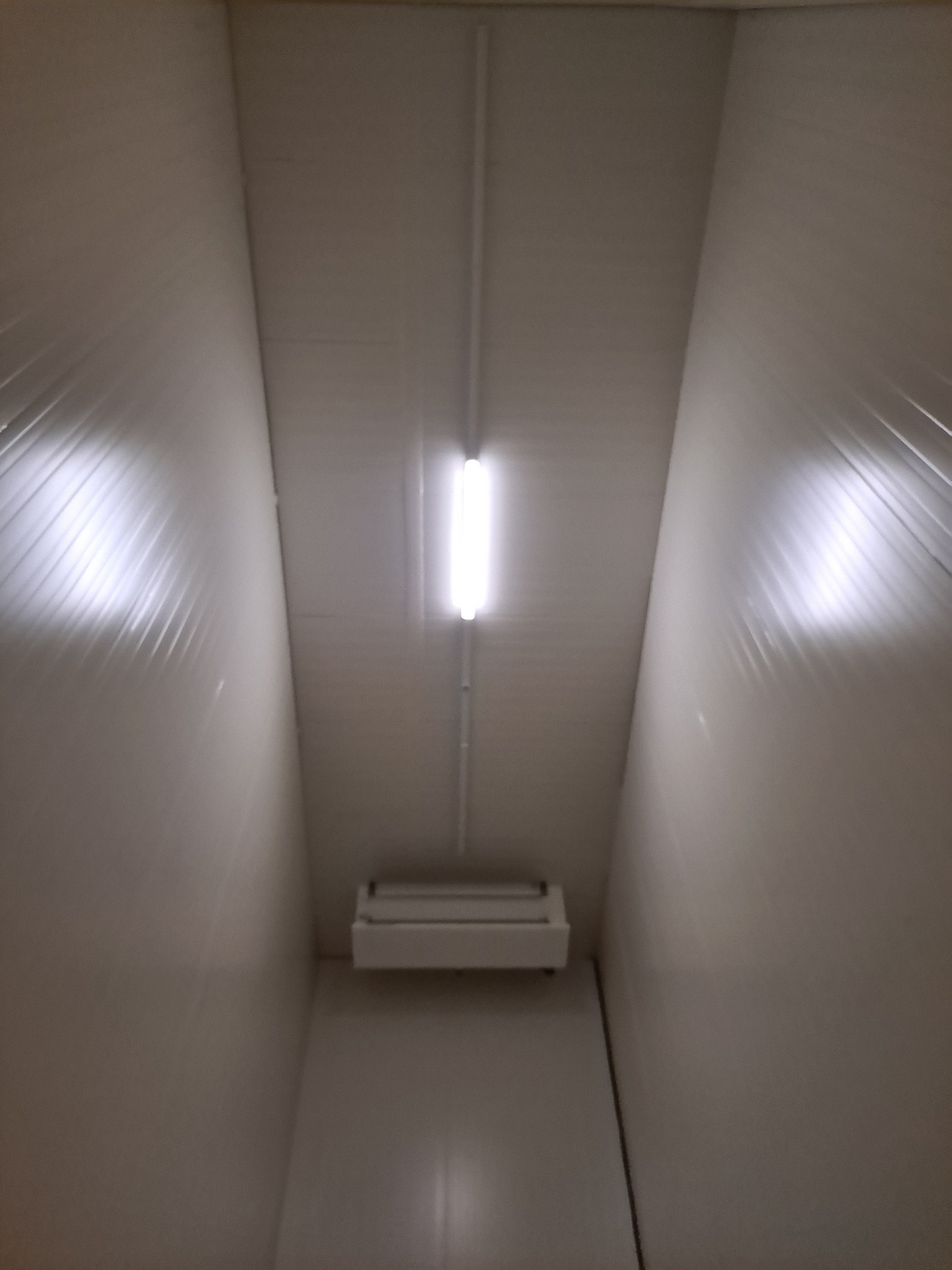 Wnętrze korytarza z białymi panelami ściennymi, oświetlone centralnie umieszczoną lampą fluorescencyjną i dodatkowym urządzeniem oświetleniowym pod sufitem.