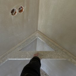 Budnarz Radosław Bednarz - Świeżo nałożona warstwa gładzi gipsowej na suficie, widoczne otwory na instalacje elektryczne i ręka osoby sprawdzającej jakość wykończenia narożnika sufitu.