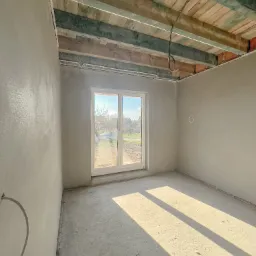 Wnętrze surowego pomieszczenia po nałożeniu tynków, widoczne drewniane elementy stropu, okno balkonowe z widokiem na podwórze, promienie słoneczne padające na podłogę.