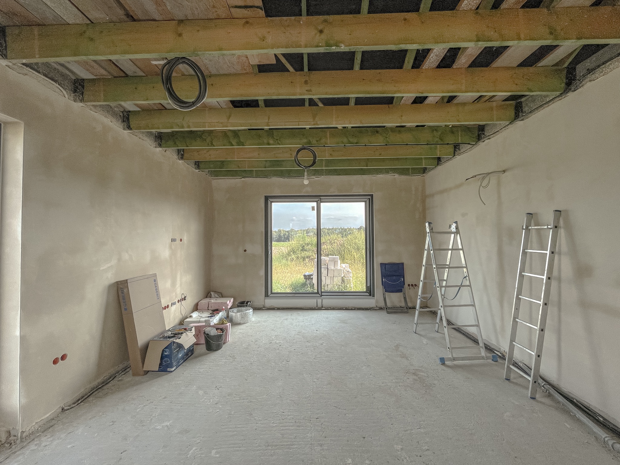 Surowe wnętrze w trakcie prac wykończeniowych; widoczne ściany po nałożeniu tynku, drewniane belki stropowe z instalacją elektryczną, drabiny aluminiowe, okno z widokiem na zewnątrz i materiały...