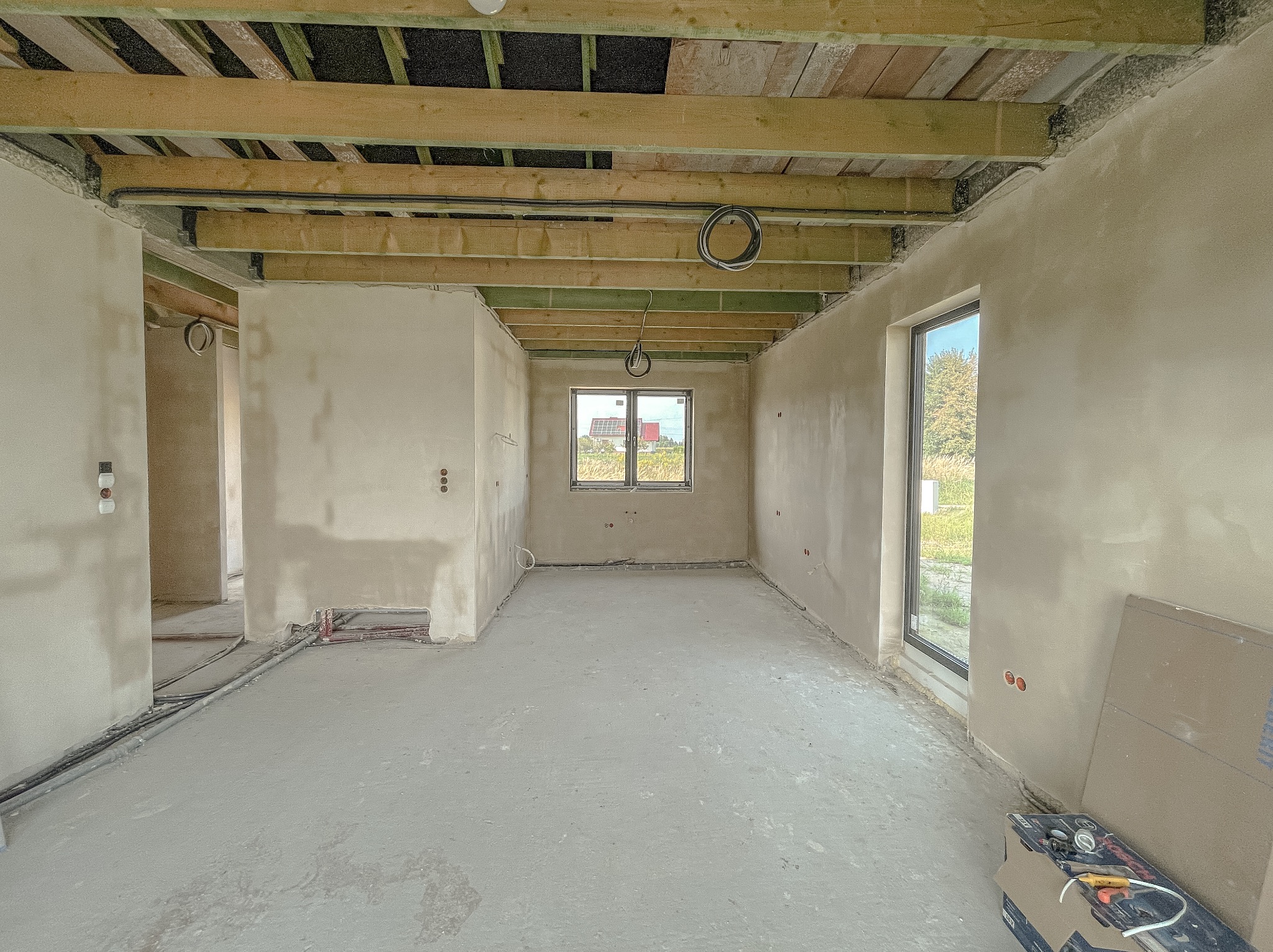 Wnętrze w stanie surowym po nałożeniu tynków, widoczne drewniane belki stropowe, instalacje elektryczne, okno z widokiem na zewnątrz oraz narzędzia na podłodze.