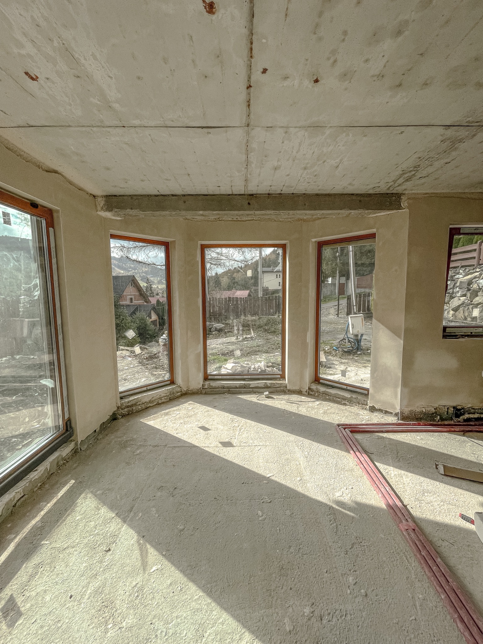 Wnętrze w stanie surowym z widocznymi oknami z brązowymi ramami, świeżo położonymi tynkami na ścianach i surowym betonowym sufitem, z promieniami słońca wpadającymi do środka i elementami...