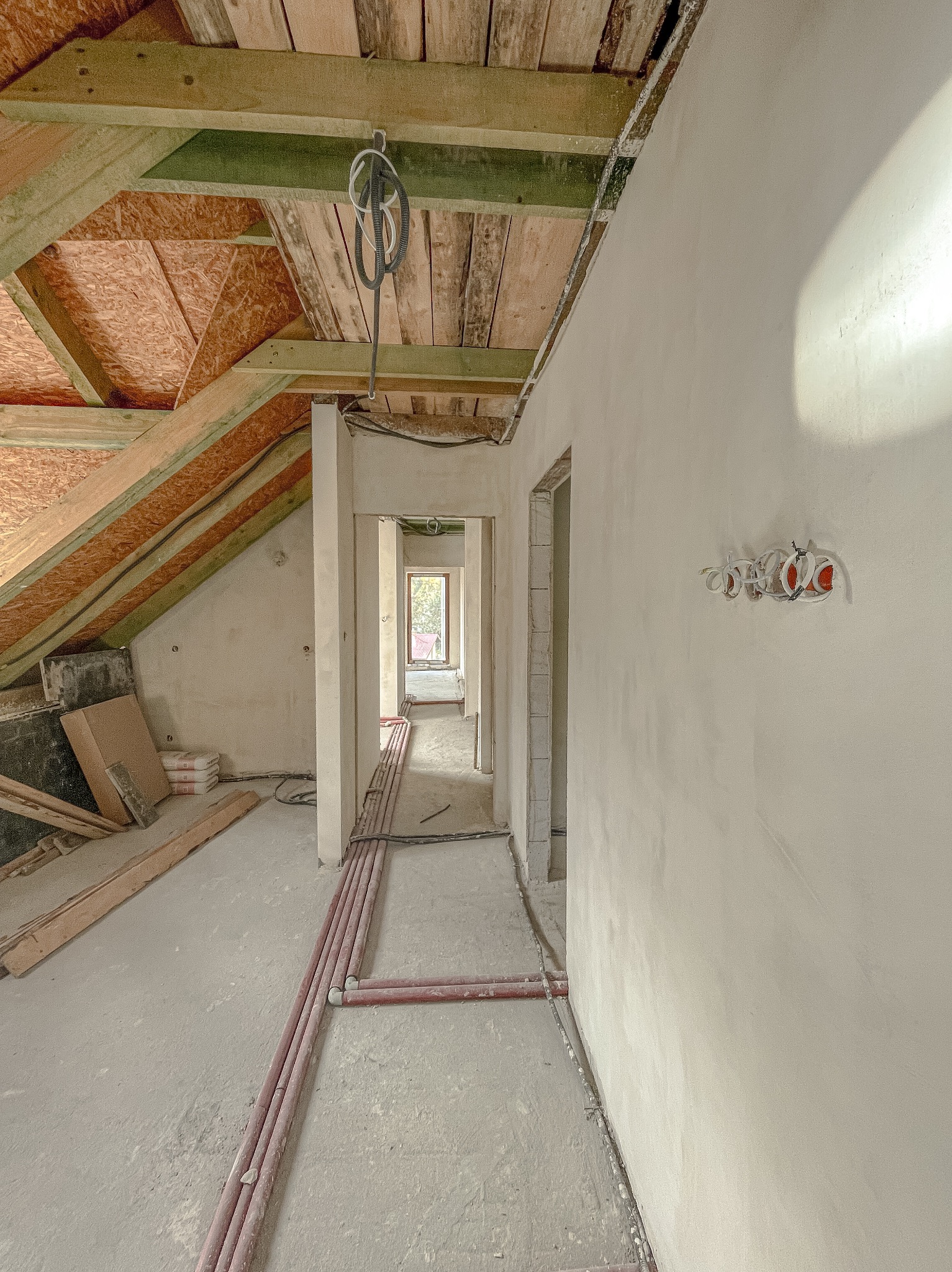 Wnętrze poddasza w trakcie prac wykończeniowych: gładkie, jasne ściany po nałożeniu tynku, widoczne instalacje elektryczne w puszkach, rury grzewcze przy podłodze oraz drewniana konstrukcja dachu...