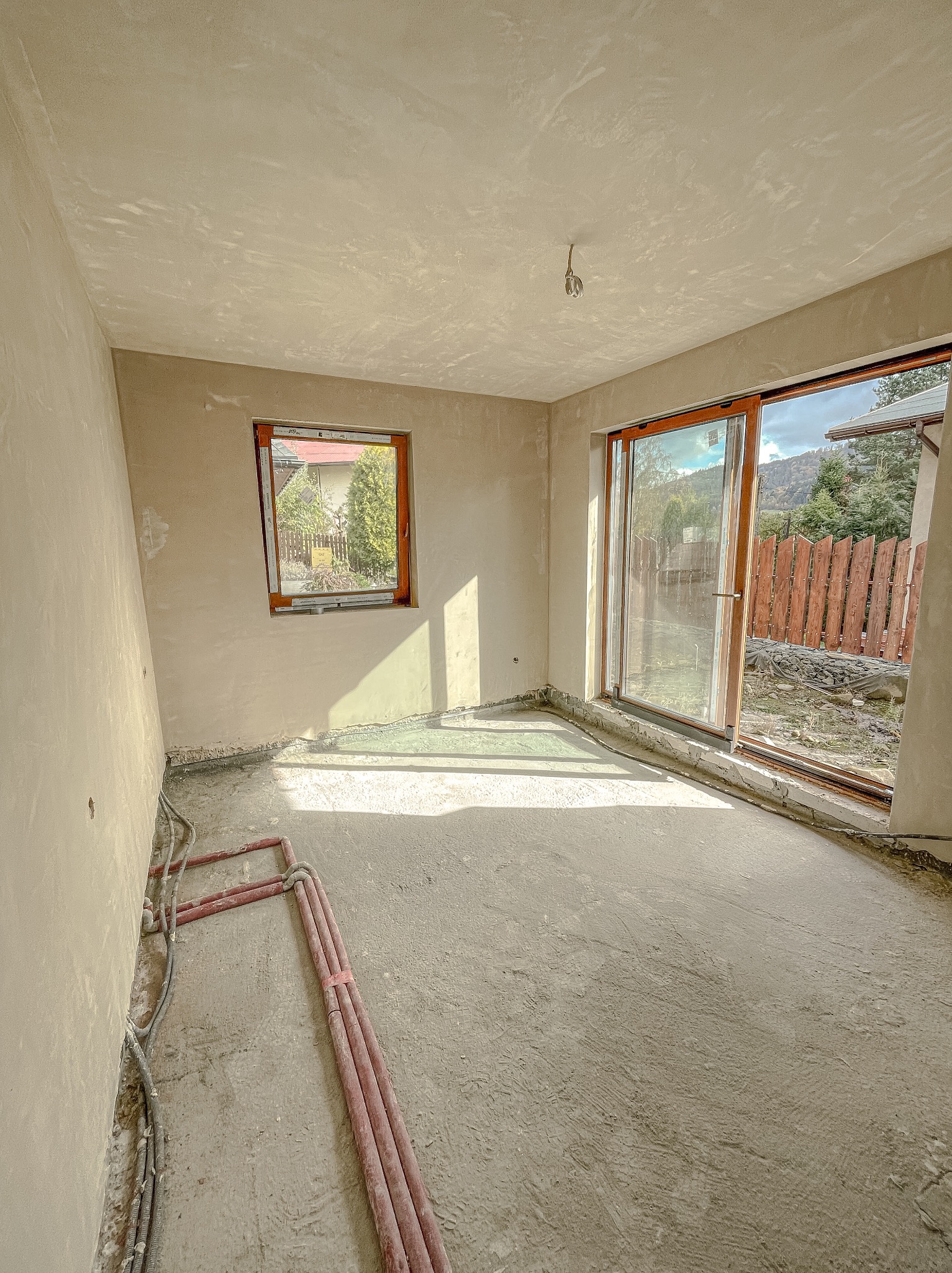 Surowe wnętrze z tynkami maszynowymi w trakcie schnięcia, widoczne rury instalacji grzewczej przy ścianie i otwarte przesuwne drzwi balkonowe z widokiem na ogród.