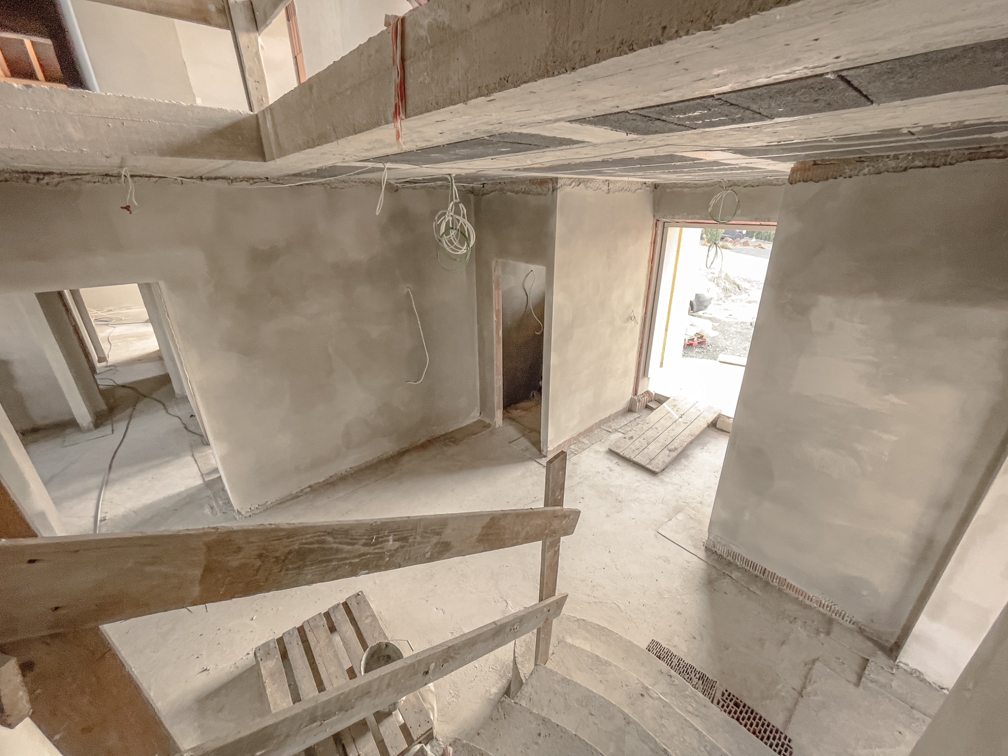 Wnętrze budynku w trakcie prac wykończeniowych, widoczne świeżo otynkowane ściany, drewniane schody z prowizoryczną barierką i wystające przewody elektryczne.