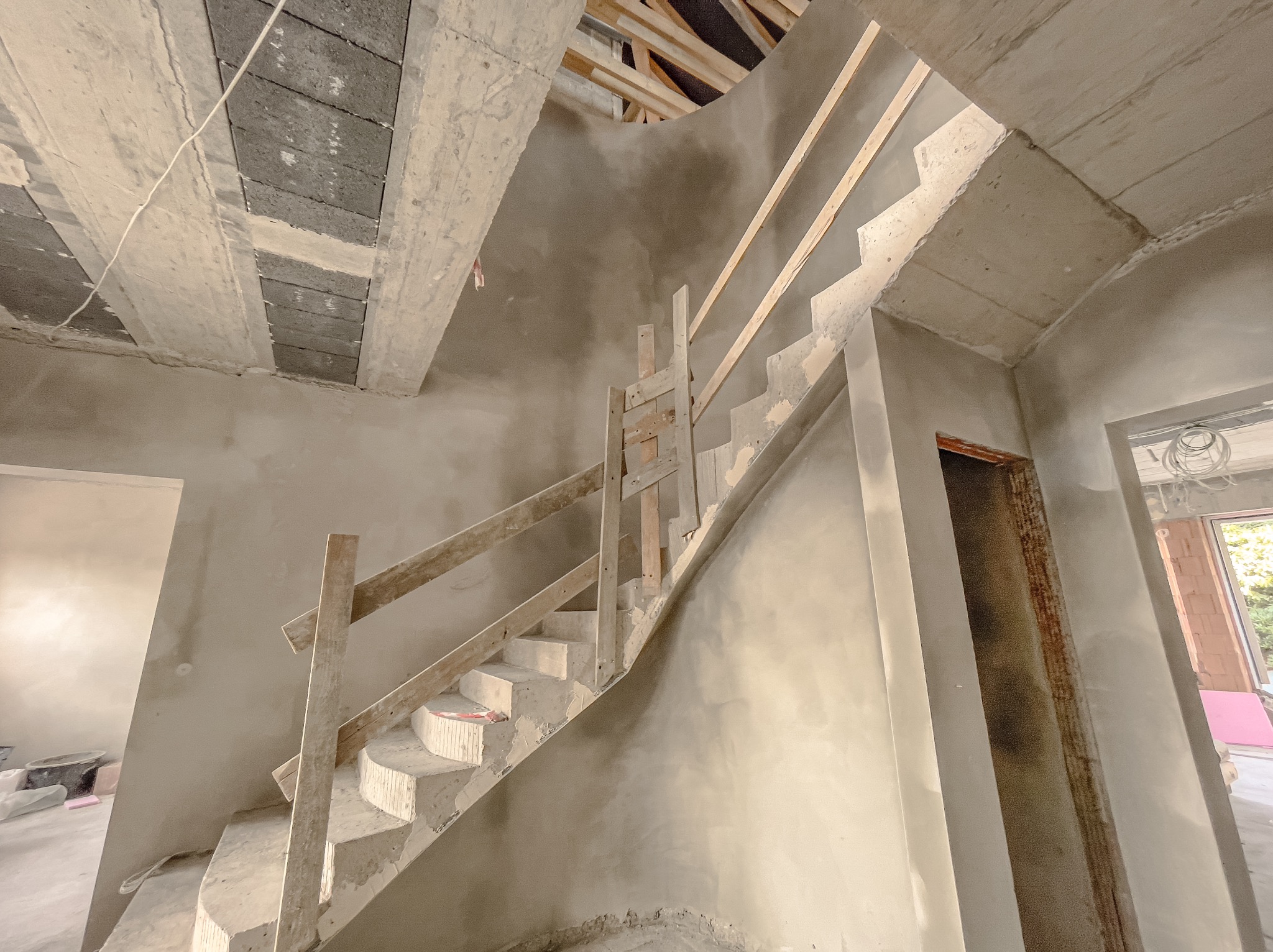 Surowe, betonowe schody z drewnianą balustradą w trakcie prac tynkarskich; widoczne ślady zaprawy na ścianach i schodach, sufit z pustaków szalunkowych.
