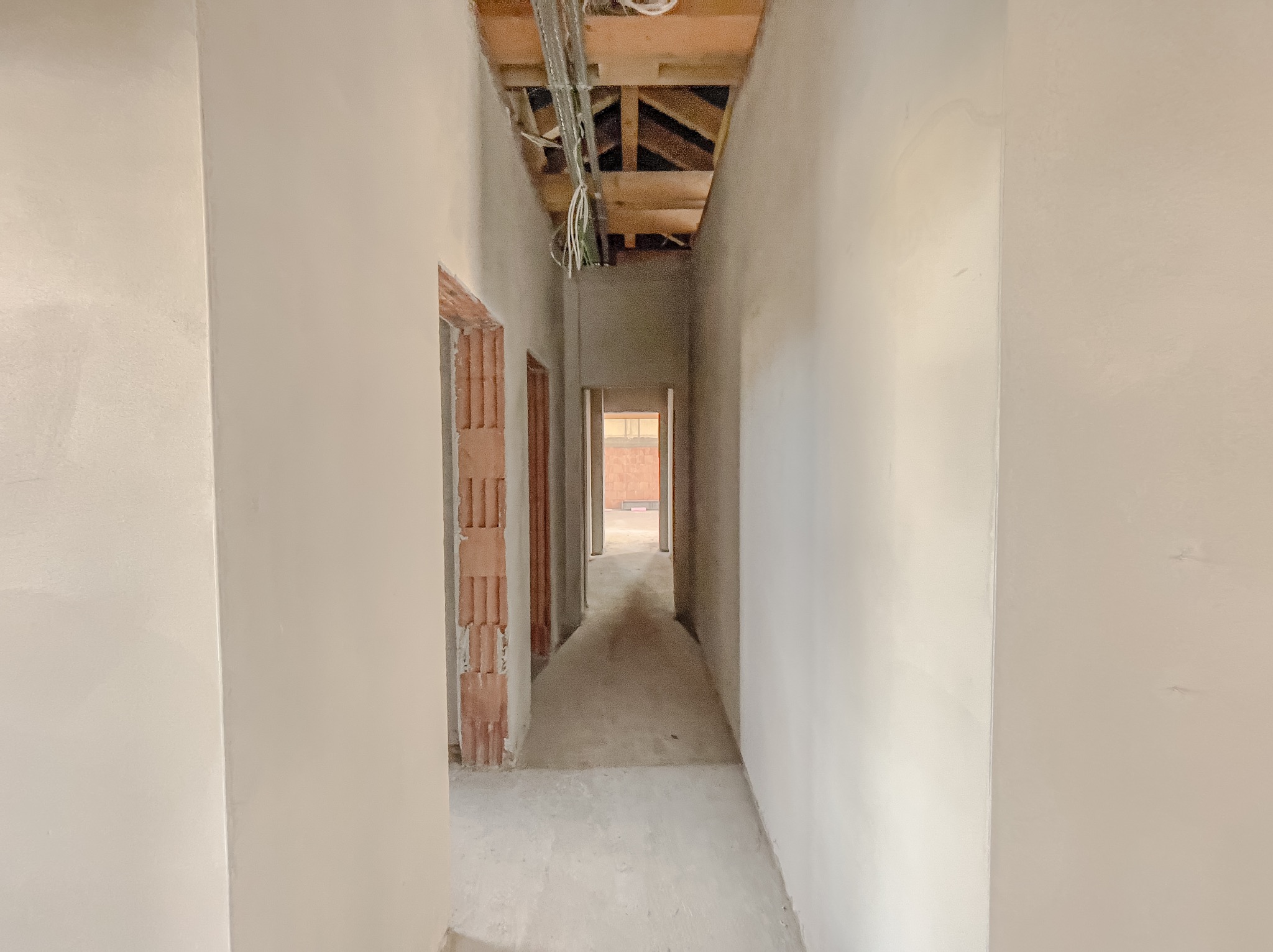Korytarz w trakcie prac wykończeniowych; ściany pokryte świeżym, jasnym tynkiem, widoczne instalacje pod sufitem, surowy stan futryn drzwiowych.
