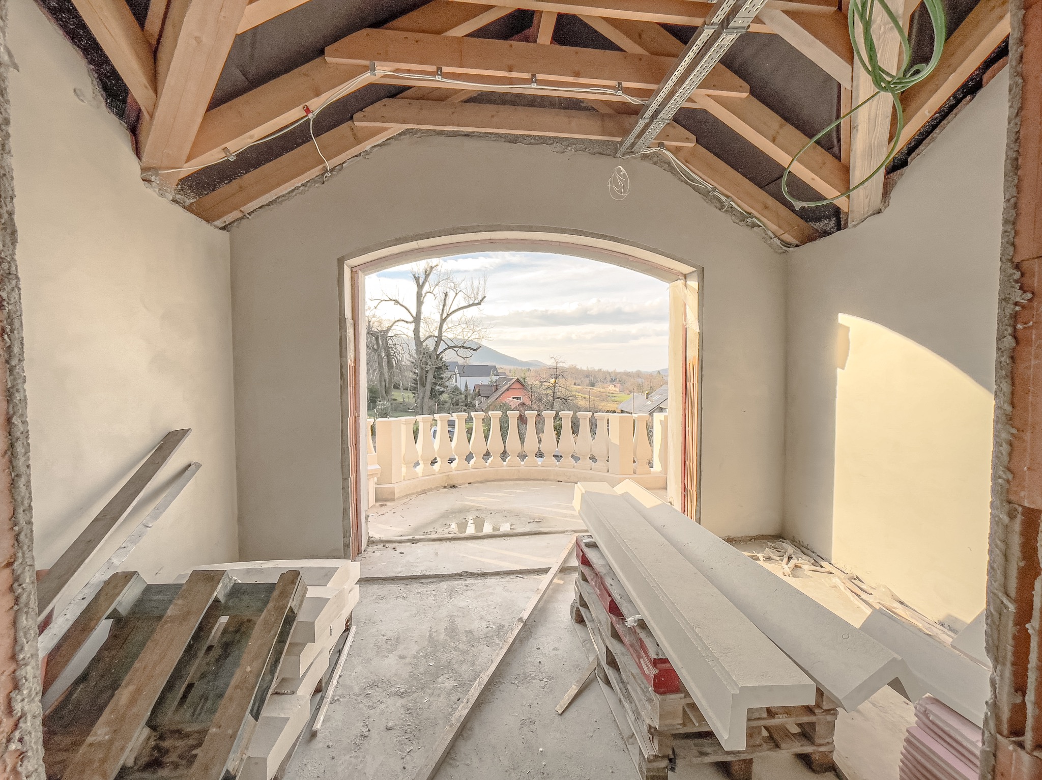 Wnętrze w trakcie wykańczania, ściany pokryte świeżym jasnym tynkiem, widoczne drewniane elementy konstrukcji dachu, przez łukowate przejście widok na balkon z balustradą i krajobraz.